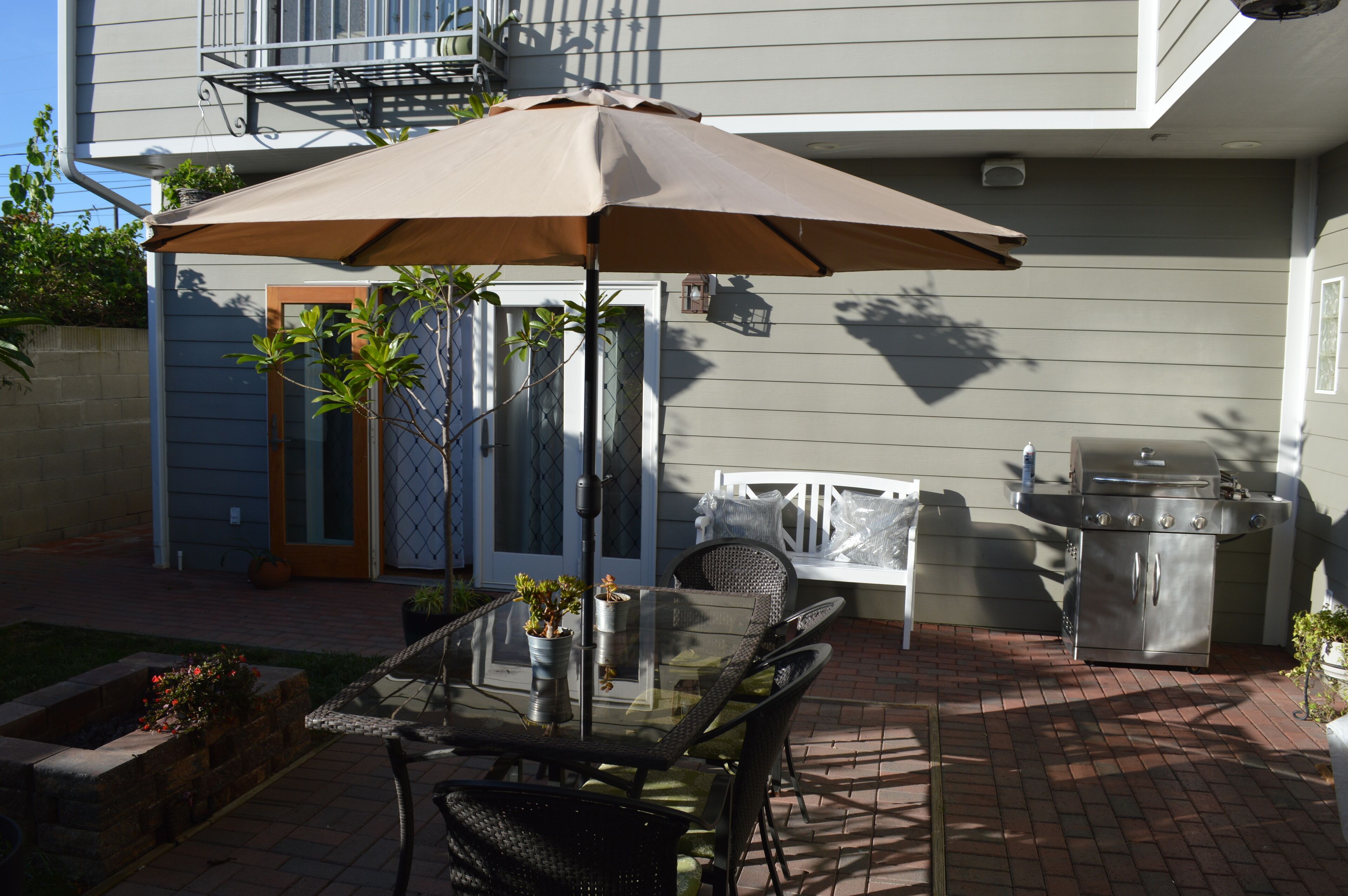 An outdoor patio with a glass table, chairs, a brown umbrella, and a barbecue grill.