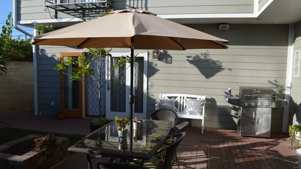 An outdoor patio with a glass table, chairs, a brown umbrella, and a barbecue grill.