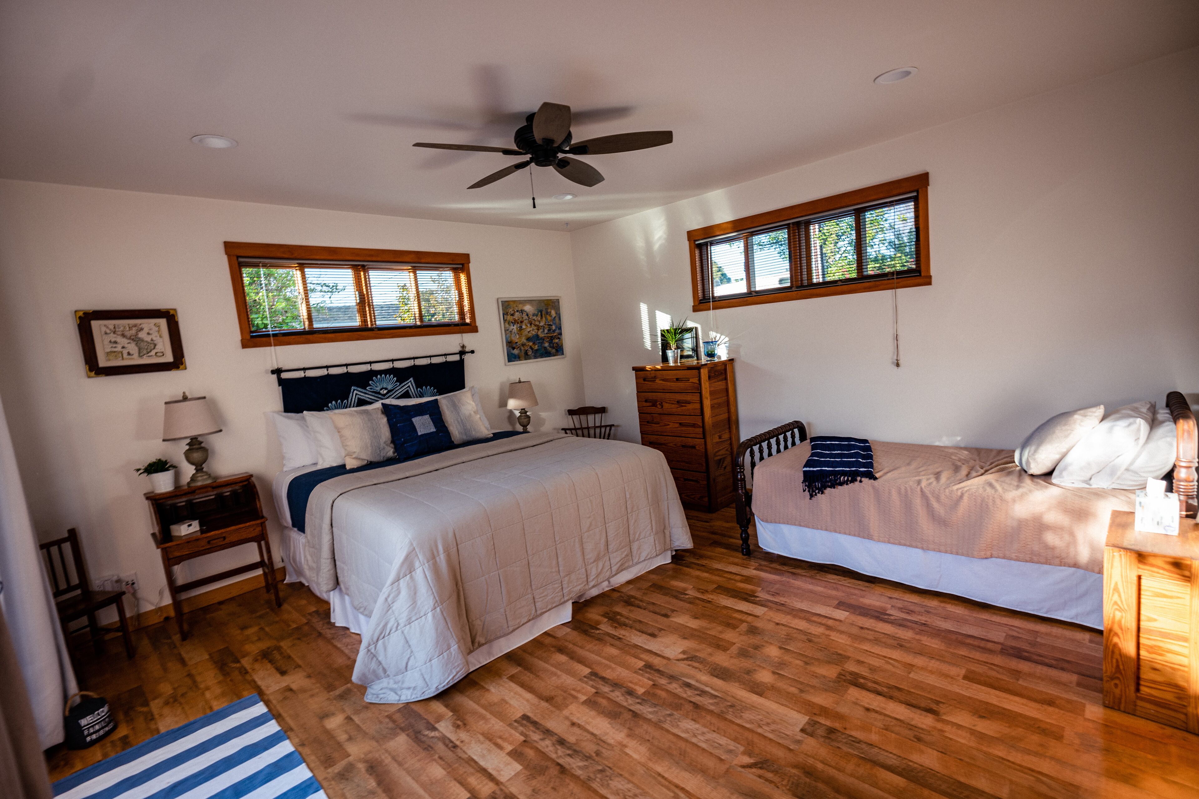 A bedroom with two beds, wooden flooring, a ceiling fan, and windows with blinds.
