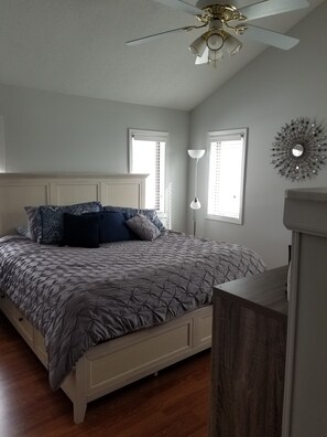 A bedroom with a bed, a ceiling fan, two windows with blinds, a floor lamp, and a decorative wall piece.