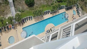 Una gran piscina al aire libre con múltiples carriles, rodeada de sillas de descanso y tumbonas, y un puente que conecta con una zona cercana.