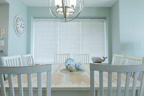 A dining area with a wooden table, white chairs, a chandelier, a clock, and a window with blinds. - Enjoy our beachfront property! (North Myrtle Beach)