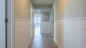 Un pasillo con paneles blancos y suelo de madera que conduce a una habitación con un ventanal y un adorno en la pared.