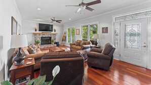 Ein geräumiges Wohnzimmer mit Kamin, Ledersofas, einem Flachbildfernseher, Deckenventilatoren und großen Fenstern.