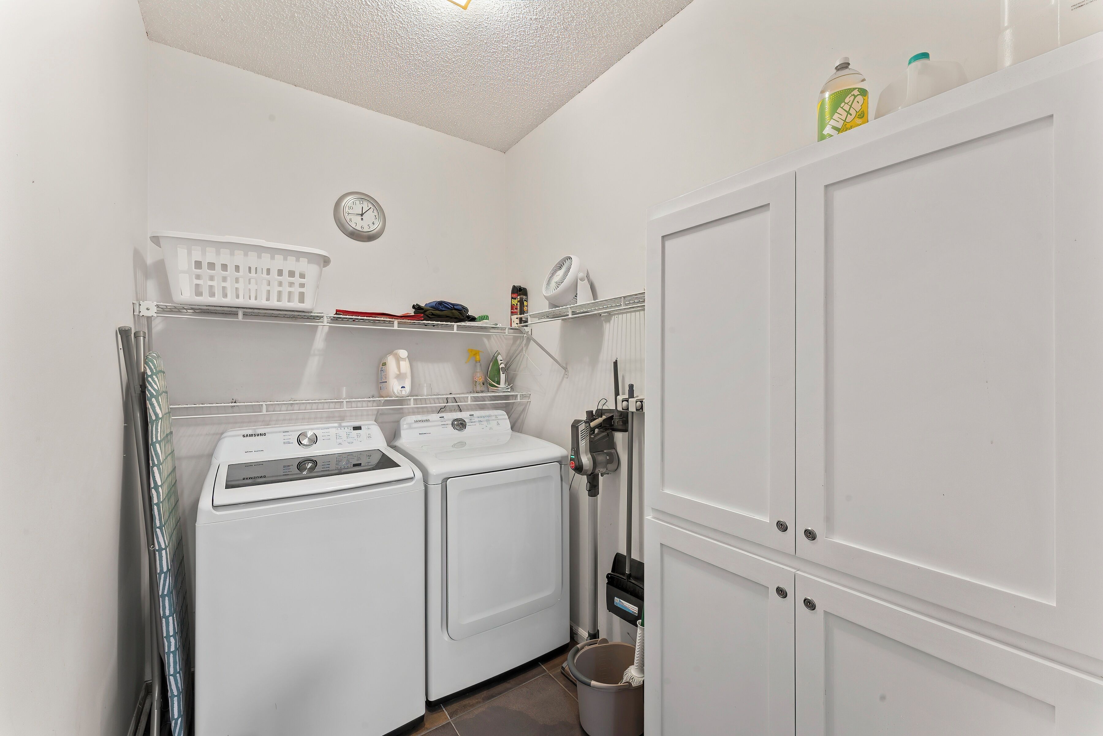 A laundry room with two white washing machines, a drying rack, a basket, and a clock on the wall.