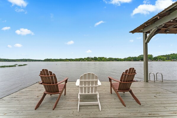 Una terraza de madera con dos sillas Adirondack marrones y una silla Adirondack blanca, orientadas hacia un lago tranquilo.