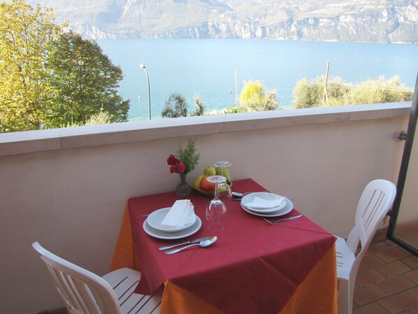 Une table dressée sur un balcon, avec des assiettes, des couverts et un vase de fleurs, d’où l’on peut admirer un lac avec des montagnes en arrière-plan.