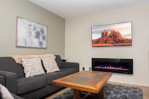 Ein Wohnzimmer mit einer Couch, einem Couchtisch, einem Kamin und zwei an der Wand befestigten Fernsehern, die unterschiedliche Szenen zeigen.