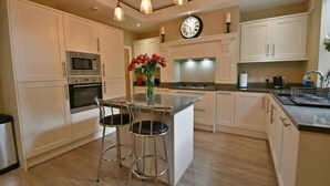 Une cuisine moderne avec des armoires blanches, des appareils en acier inoxydable, un îlot central doté de tabourets, des luminaires suspendus et une horloge au mur.