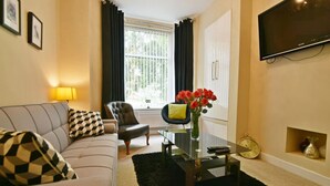 Un salon comprenant un canapé, un fauteuil et une table basse. On y trouve une fenêtre avec des rideaux, un téléviseur fixé au mur et un vase rempli de fleurs sur la table basse.