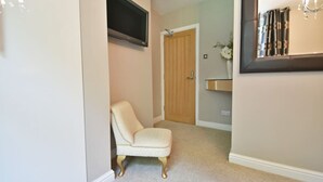 Un couloir avec un tapis beige, un fauteuil de couleur crème, une porte en bois, un téléviseur fixé au mur et un miroir avec un cadre décoratif.