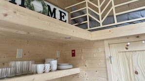 A wooden interior with a loft bed, shelves with white mugs and glasses, and a 'HOME' sign.