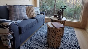 A cozy cabin interior with a blue sofa, patterned pillows, a woven rug, and a view of a forest through a large window.