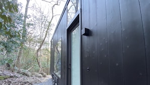 A dark wooden cabin with a large window, surrounded by trees and a gravel path.