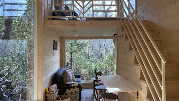 A wooden cabin interior with a loft bedroom, a dining area, and a view of the forest.