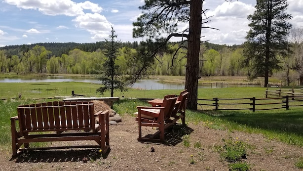 A wooden bench and chairs by a lake with a fenced area and a forested hill in the background.