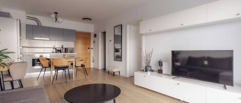 A modern living room with a flat-screen TV, a dining area with chairs, a kitchen with cabinets, and a wooden floor.