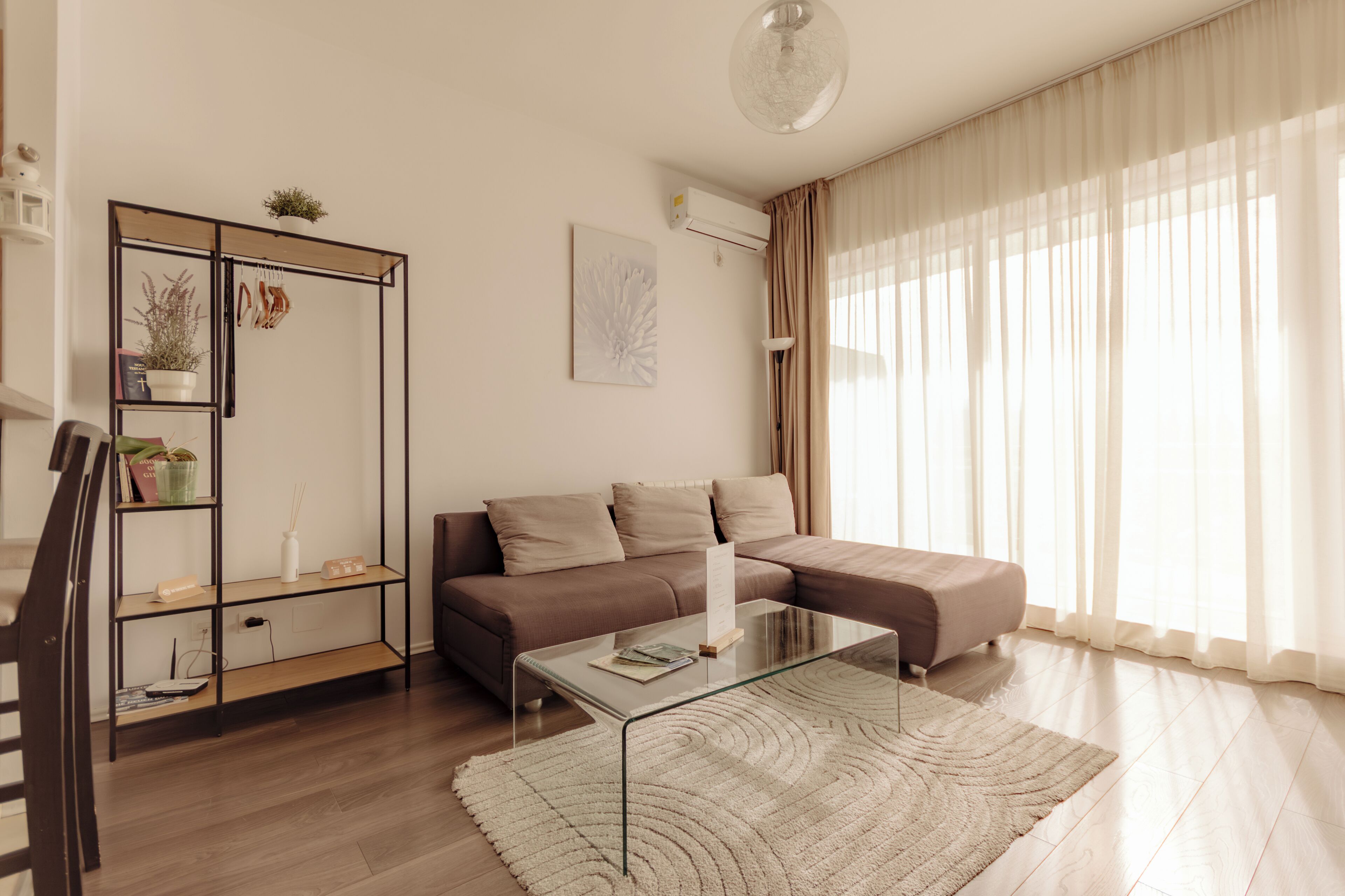 A modern living room with a glass coffee table, a brown sectional sofa, a glass-top coffee table, a white rug, and a black metal shelving unit with plants.