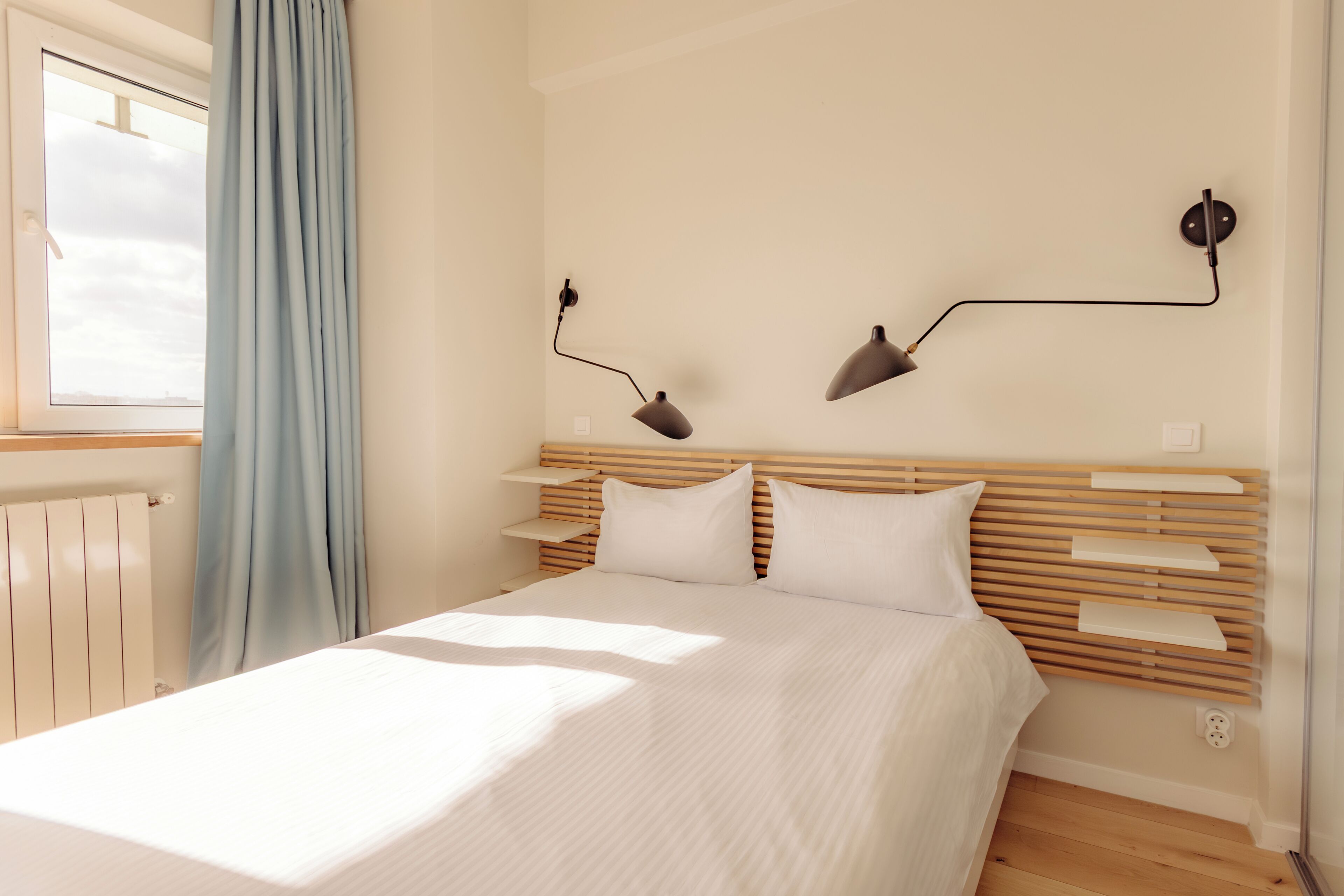 A bedroom with a wooden headboard, two bedside lamps, a window with curtains, and a radiator.