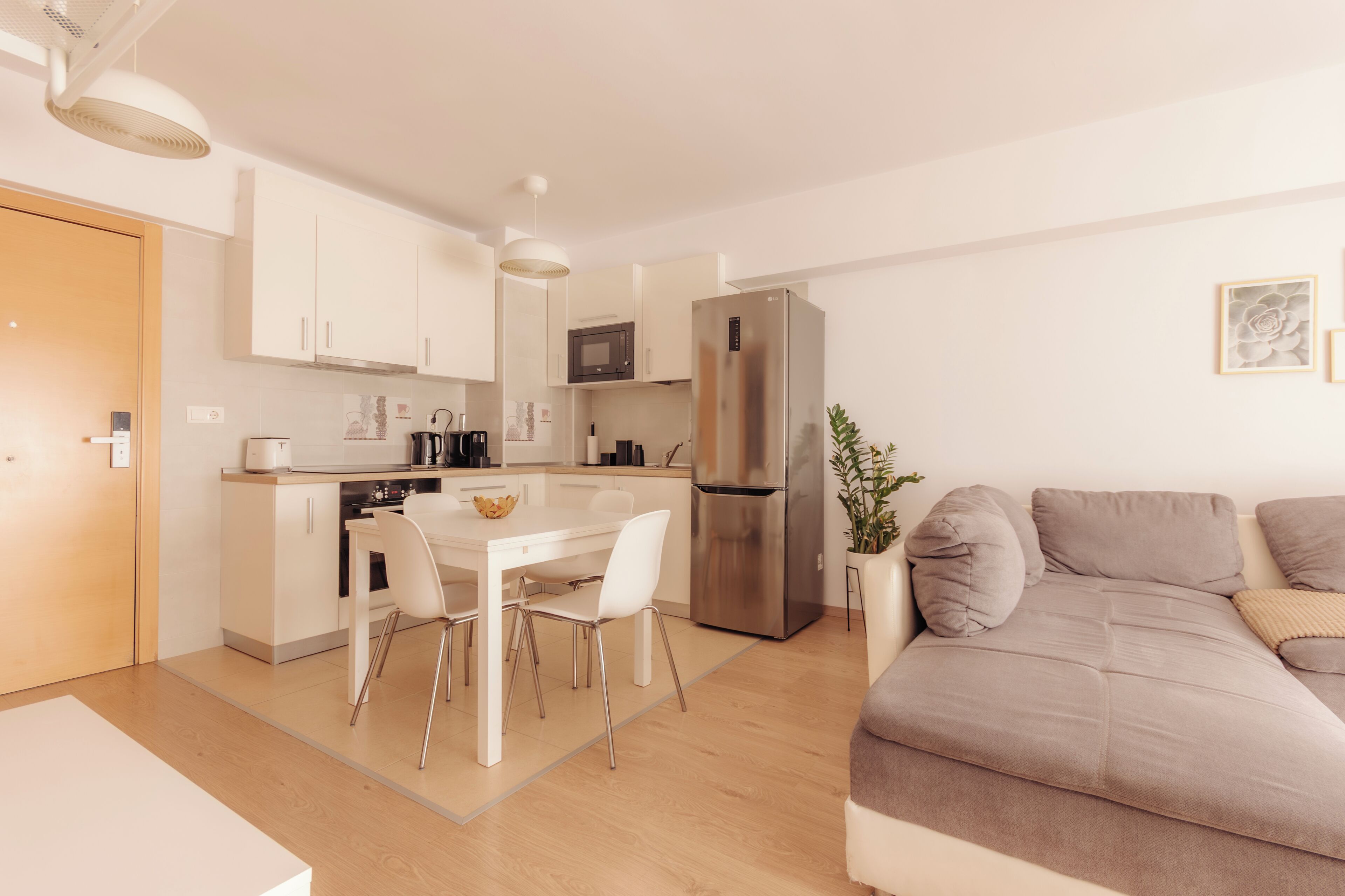 A modern kitchen with white cabinets, a stainless steel refrigerator, a dining table with chairs, and a beige sofa in the living area.
