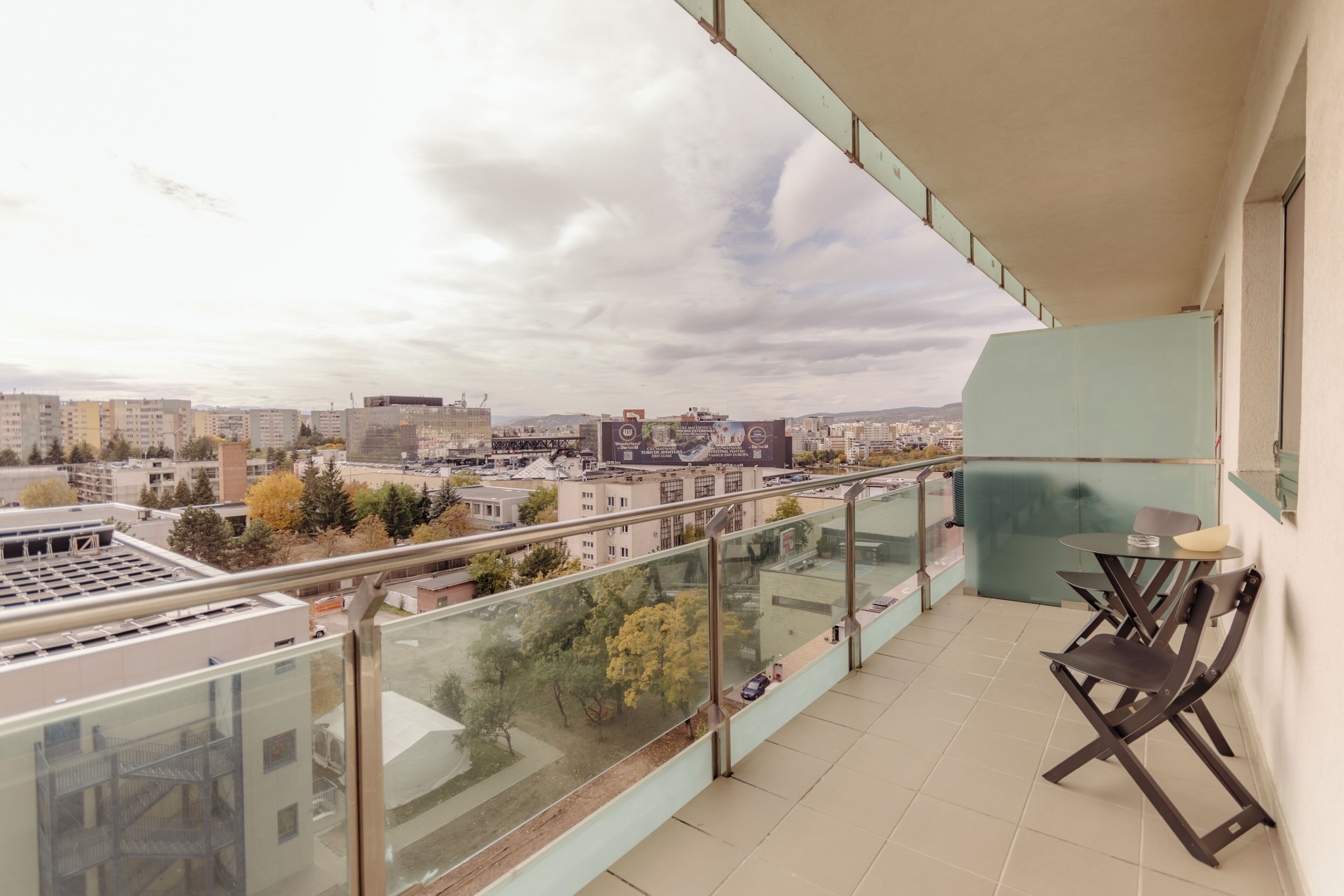 Un balcón con barandilla de vidrio, una mesa con sillas y vistas a un paisaje urbano con edificios y árboles.