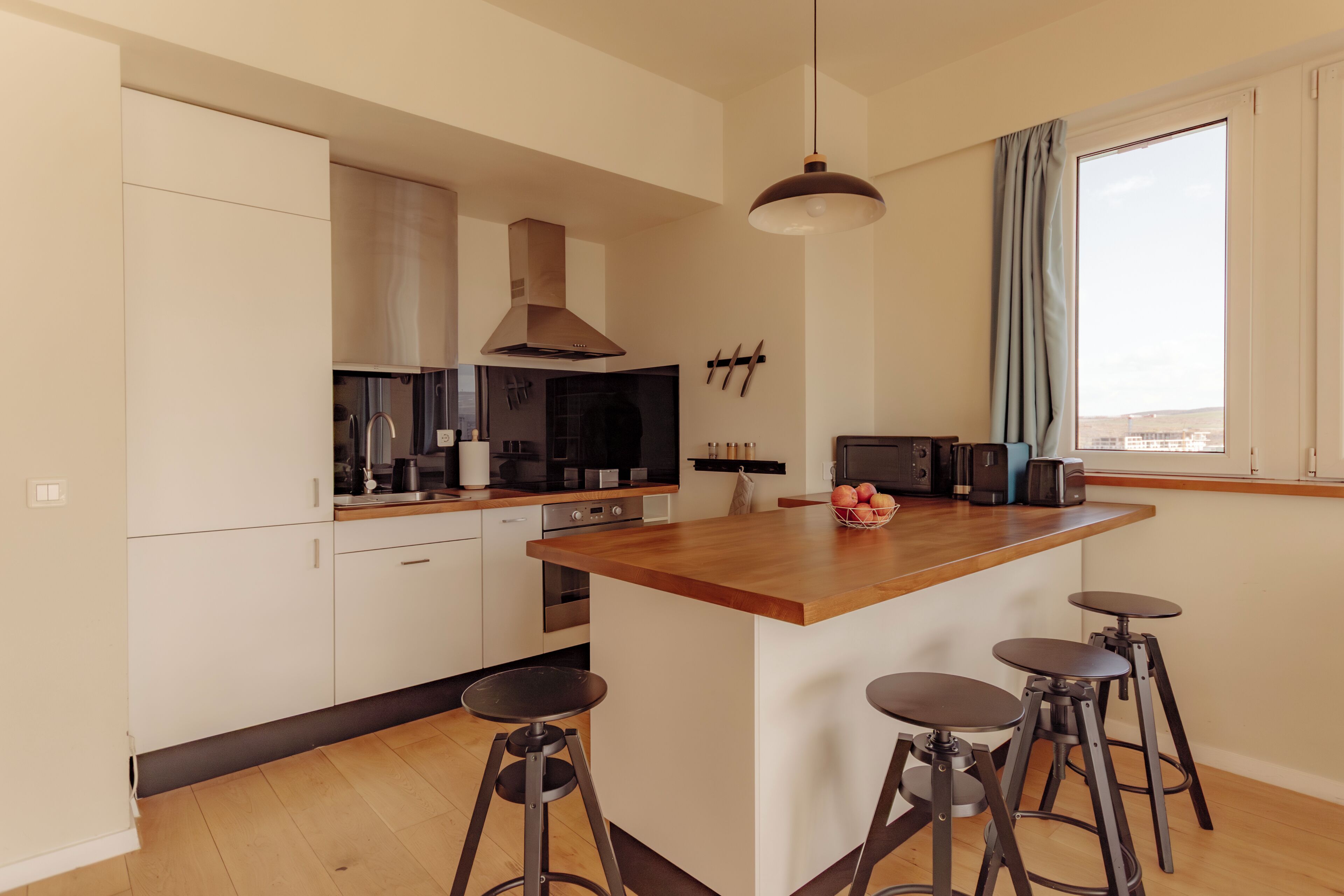 A modern kitchen with white cabinets, a wooden island, bar stools, and a large window.