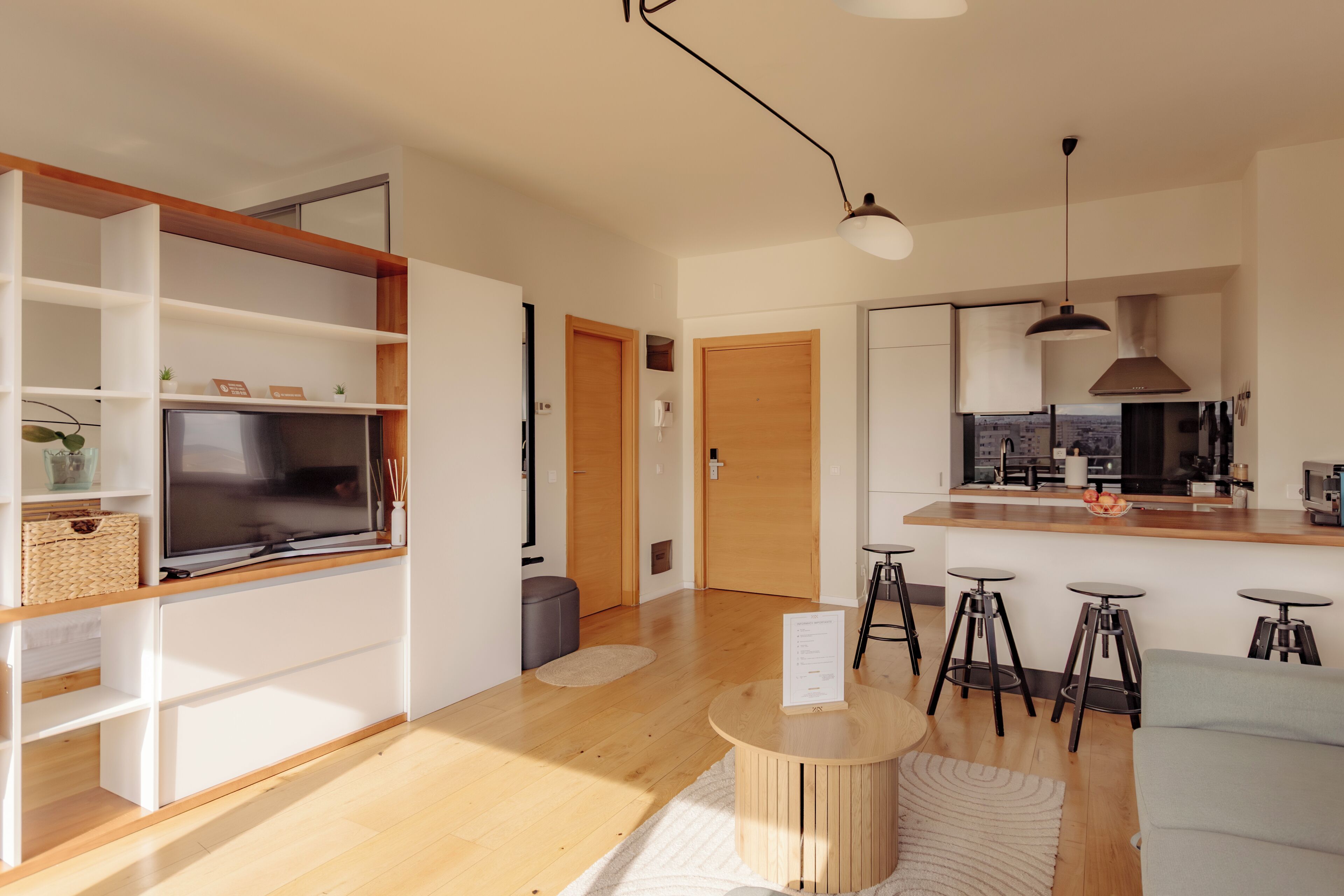 A modern living room with a TV, wooden shelves, a round coffee table, and a kitchen area with stools.