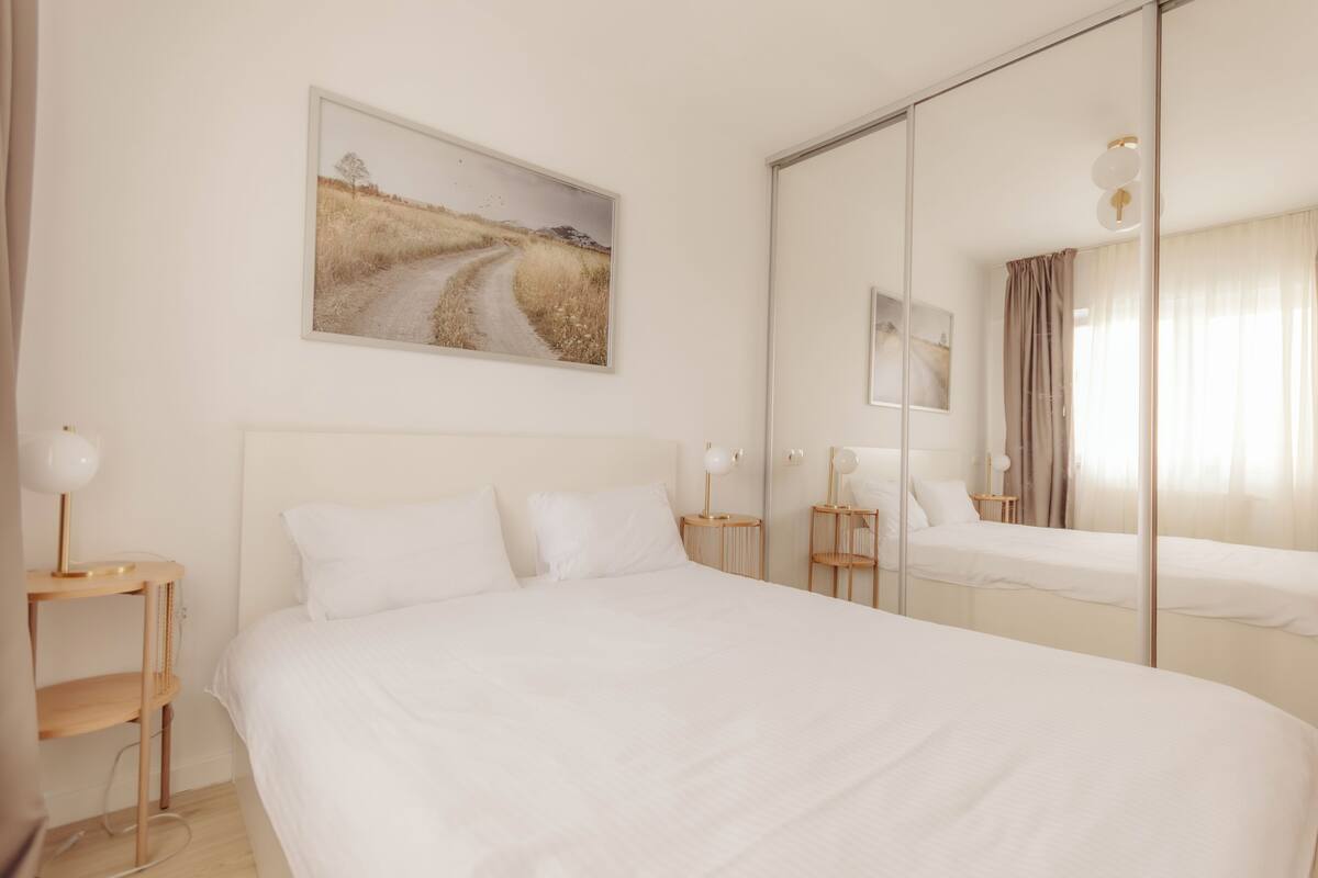 Quarto com uma cama grande, uma mesinha de cabeceira, um espelho e um quadro de uma estrada na parede.