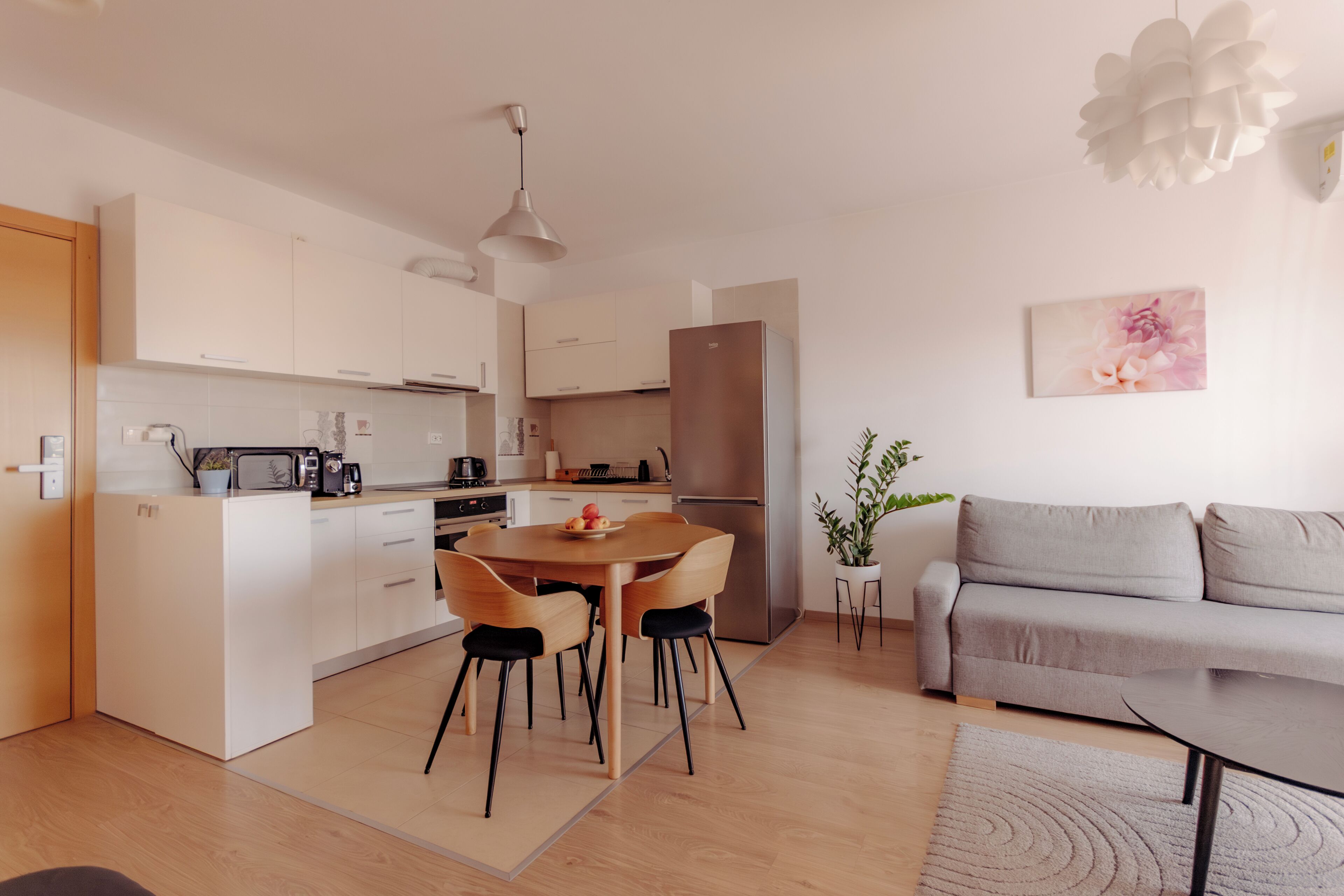 A modern kitchen with white cabinets, a dining table with chairs, a grey sofa, and a potted plant.