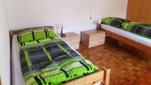 A room with two single beds, each with a green and black patterned bedspread, a wooden nightstand with a lamp, and a wooden wardrobe.