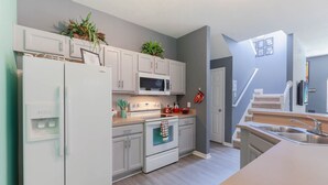 Una cocina moderna con armarios blancos, microondas integrado, estufa y refrigerador. Hay plantas sobre la encimera y una escalera que lleva a otro nivel.