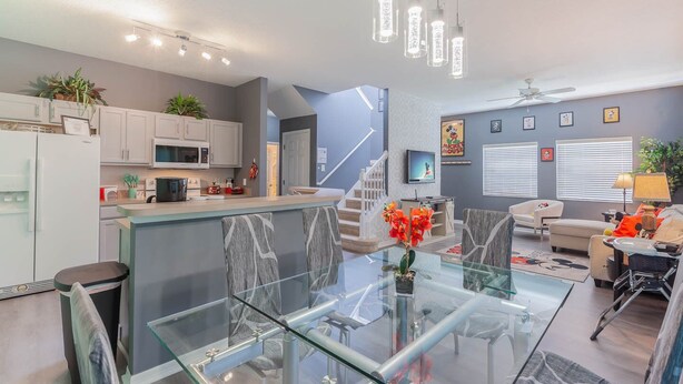 Una cocina moderna con armarios blancos, microondas y refrigerador. Hay una mesa de comedor de vidrio con sillas, una escalera que lleva a otro nivel y una sala de estar con sofá y televisor.
