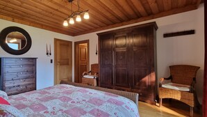 A bedroom with a wooden ceiling, a large wardrobe, a bed with patterned bedding, and a wicker chair. - Spacious, privacy and beautiful unobstructed view of the mountains and valley (Stuhlfelden)
