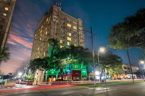 Un immeuble à plusieurs étages dont la façade est illuminée de lumière verte, avec un grand arbre devant et une rue où l’on aperçoit des traînées de lumière.