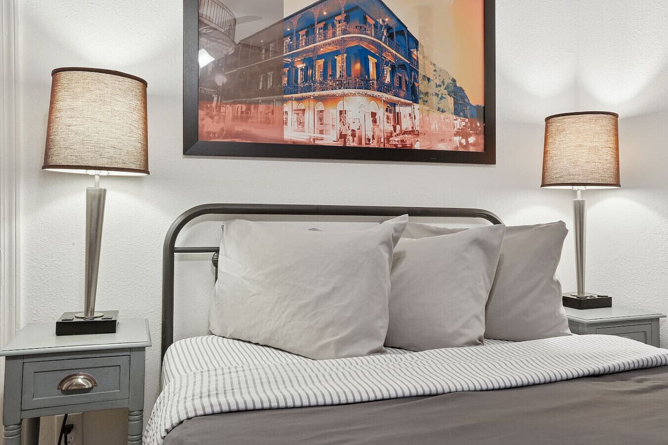 Una cama bien hecha con sábanas blancas y dos almohadas, flanqueada por dos mesitas de noche con lámparas, en una habitación con un cuadro enmarcado de una vista urbana sobre la cama.