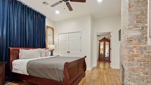 Une chambre avec un lit en bois, un ventilateur de plafond, un mur en briques et un couloir menant à une autre pièce.