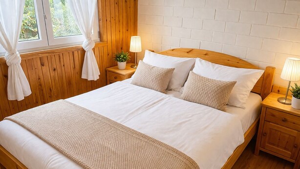 A wooden bed with white bedding, a bedside table with a lamp, and a window with curtains.
