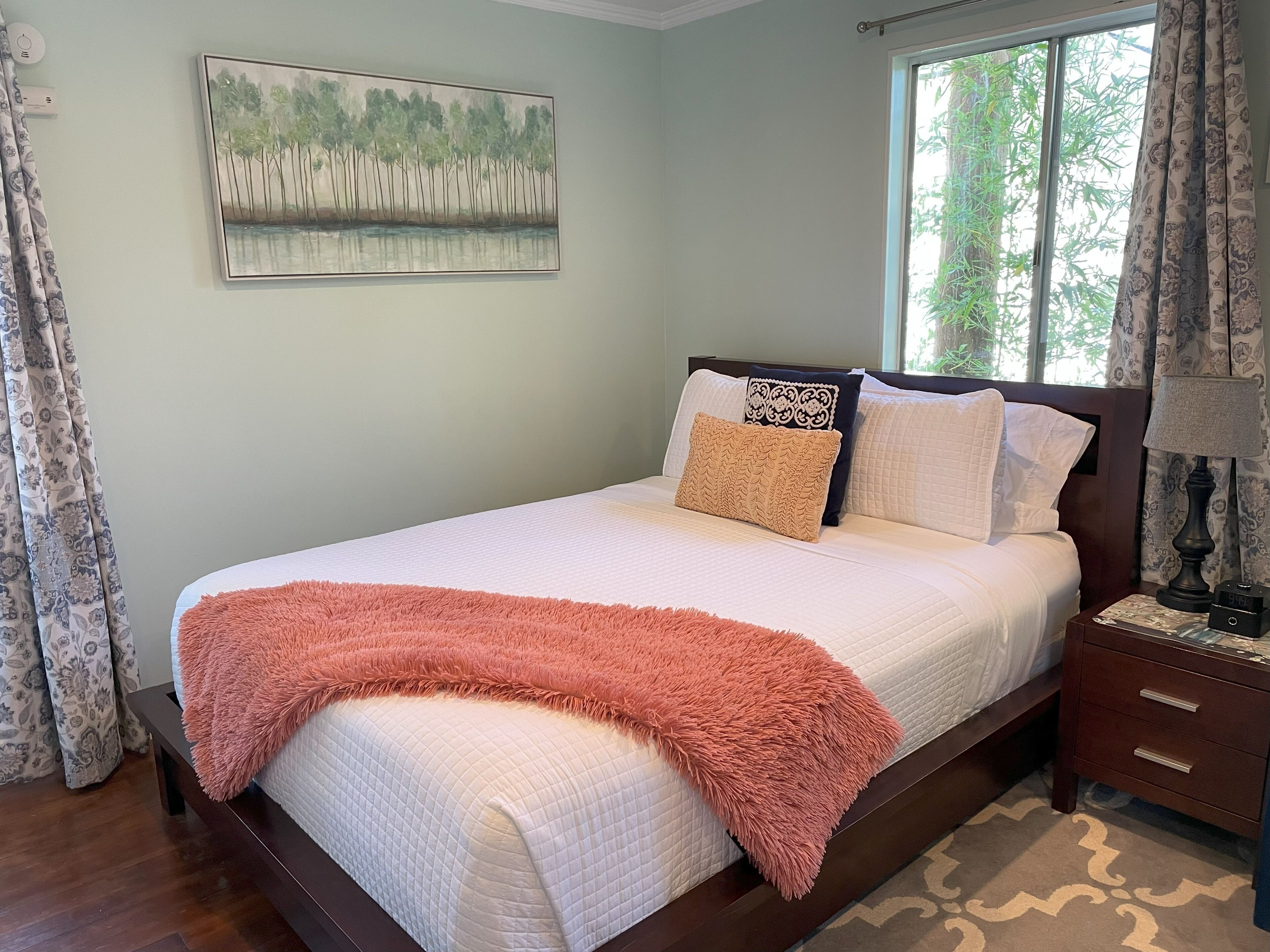 Een netjes opgemaakt bed met een wit geborduurd dekbed, twee kussens en een perzikkleurig plaid. Aan de muur hangt een schilderij van bomen en water, en er is een raam met gordijnen zichtbaar.