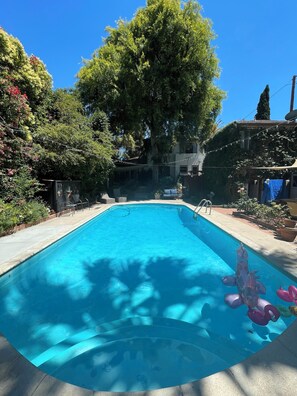 Una piscina rectangular con superficie de agua azul cristalina, rodeada por una solana de concreto. Se observan árboles y arbustos al fondo, y un edificio con techo inclinado visible parcialmente detrás de la piscina.