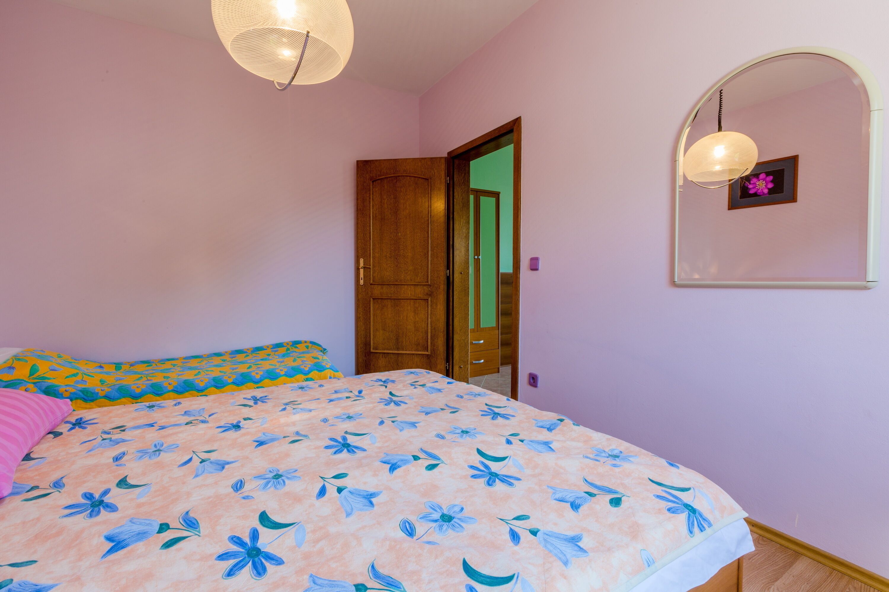 A bedroom with a floral bedspread, a wooden door, a mirror with a picture frame, and a hanging light fixture.