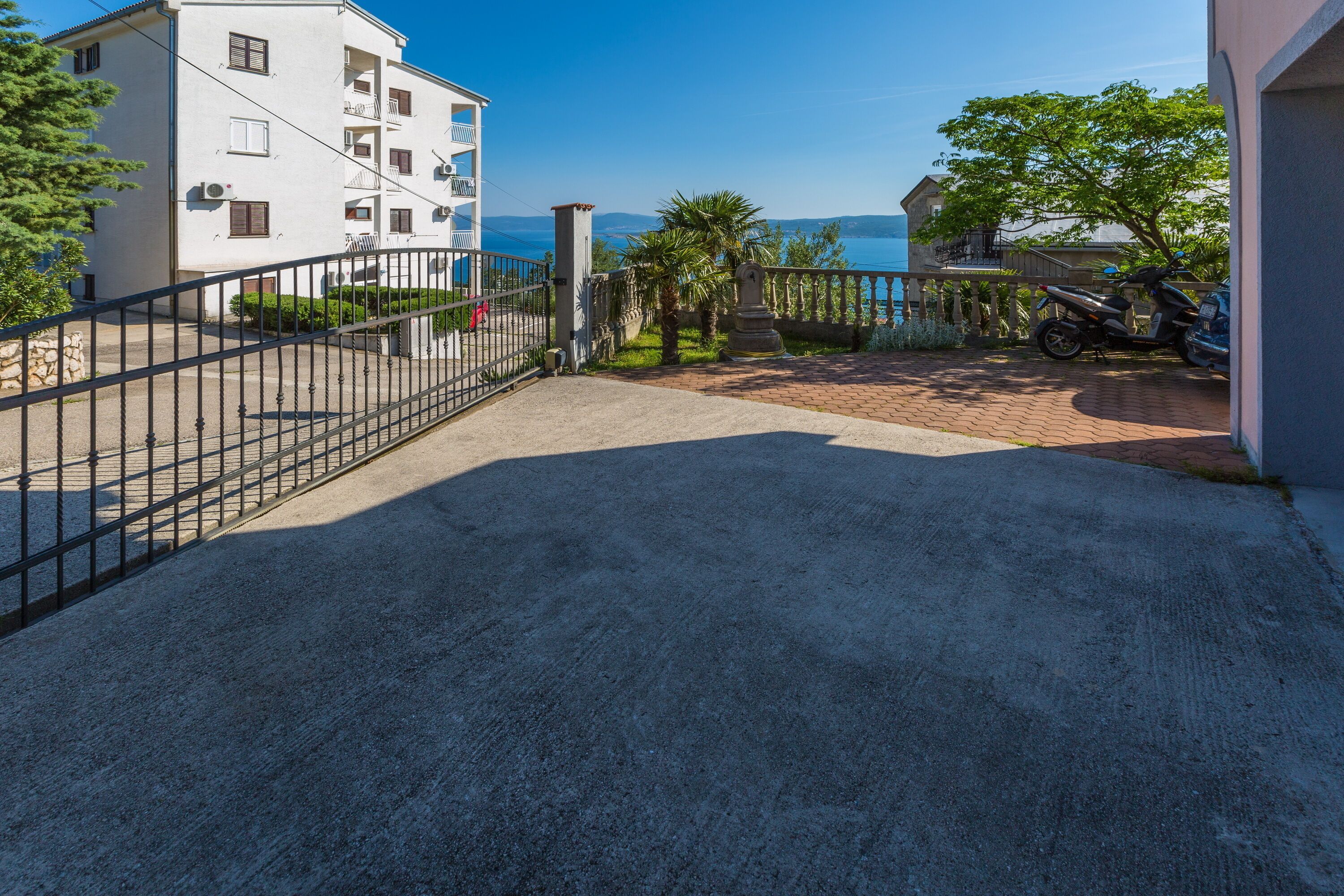 A paved driveway leading to a gated entrance with a view of a body of water and a building in the background.