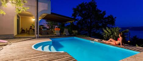 A wooden deck with a swimming pool, outdoor seating, and a view of the sea at dusk.