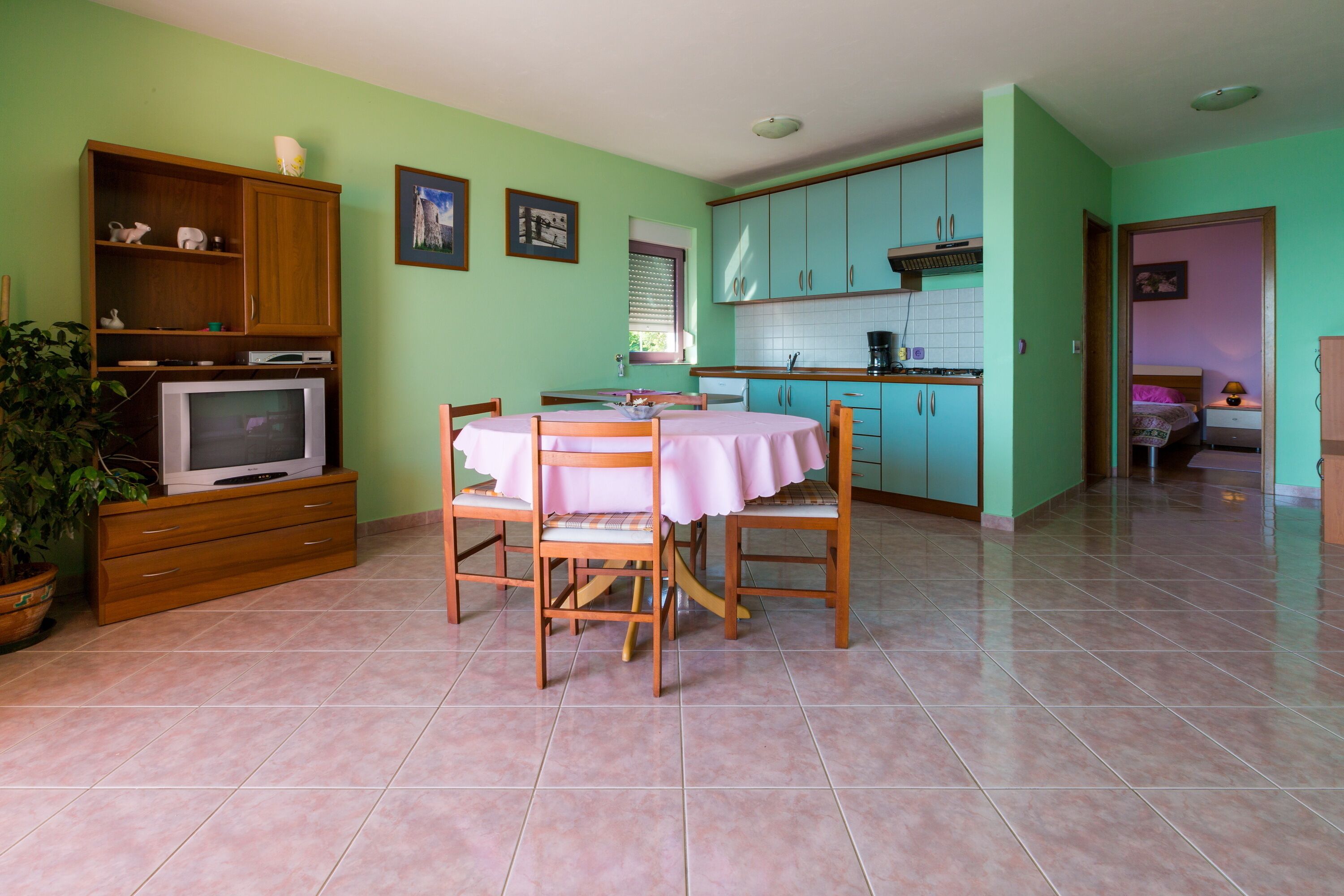 A dining area with a table and chairs, a television on a wooden cabinet, and a kitchen with teal cabinets.