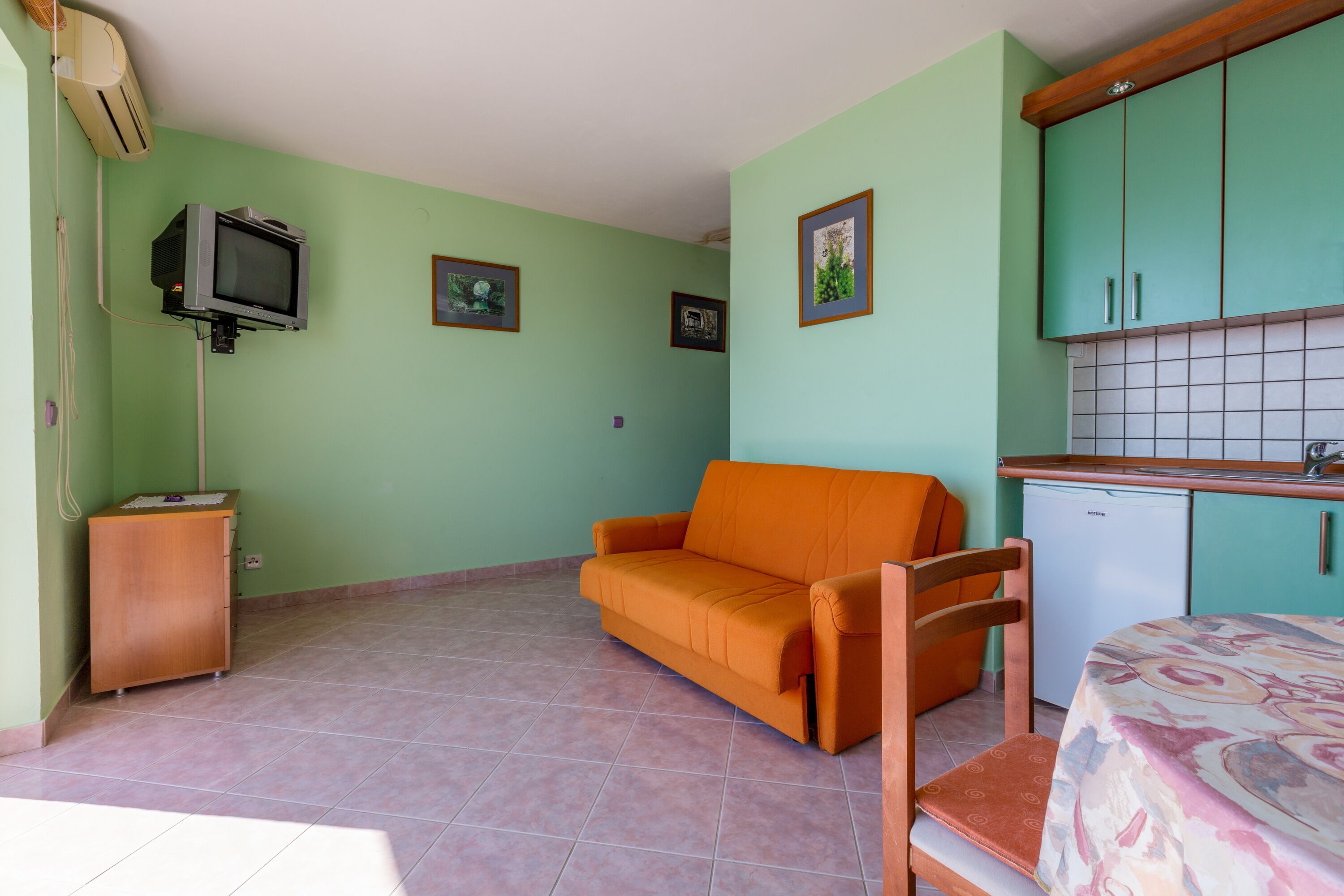 A room with a sofa, a television, a small cabinet, and a kitchenette area.