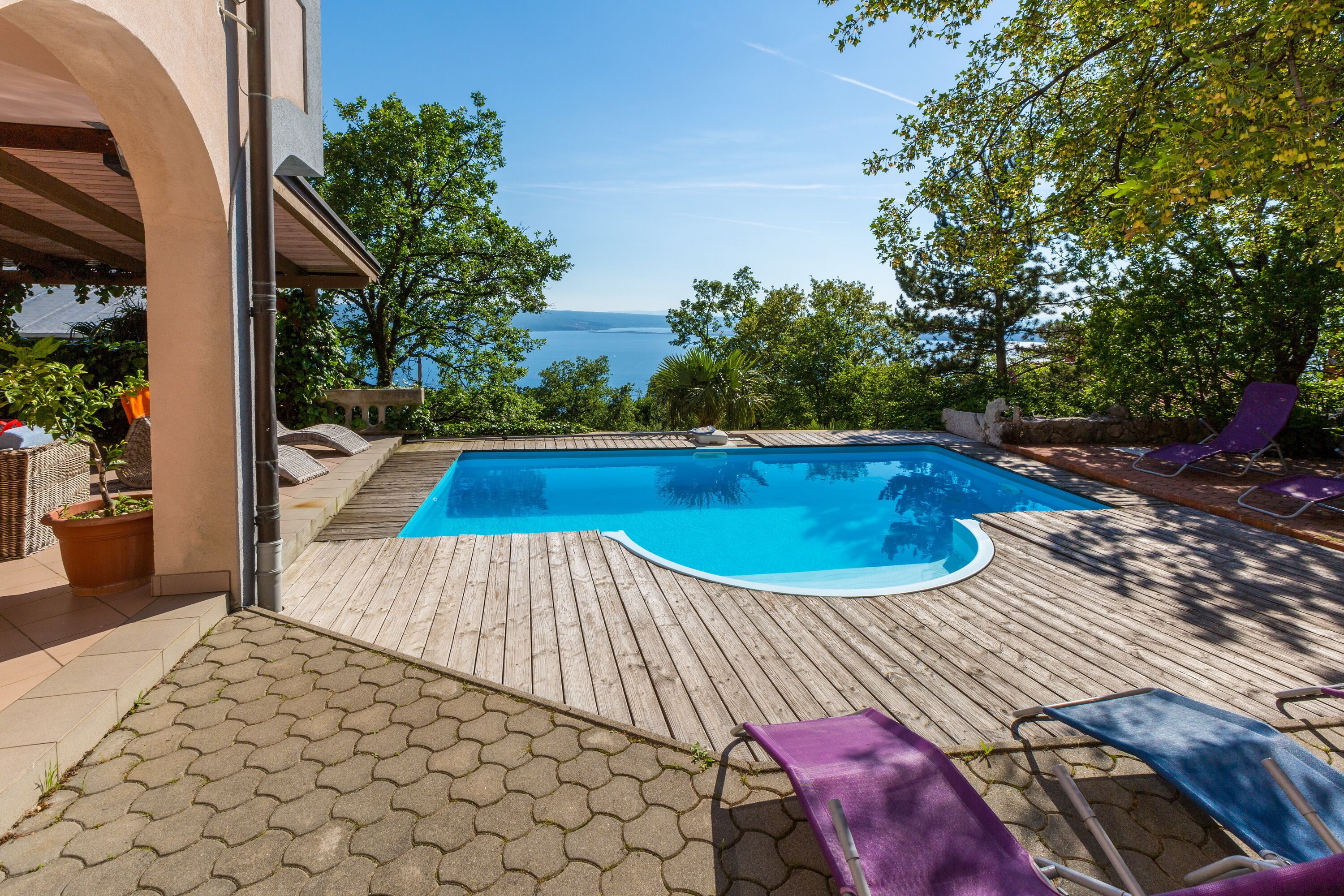 A swimming pool with a wooden deck, lounge chairs, and a view of the sea.