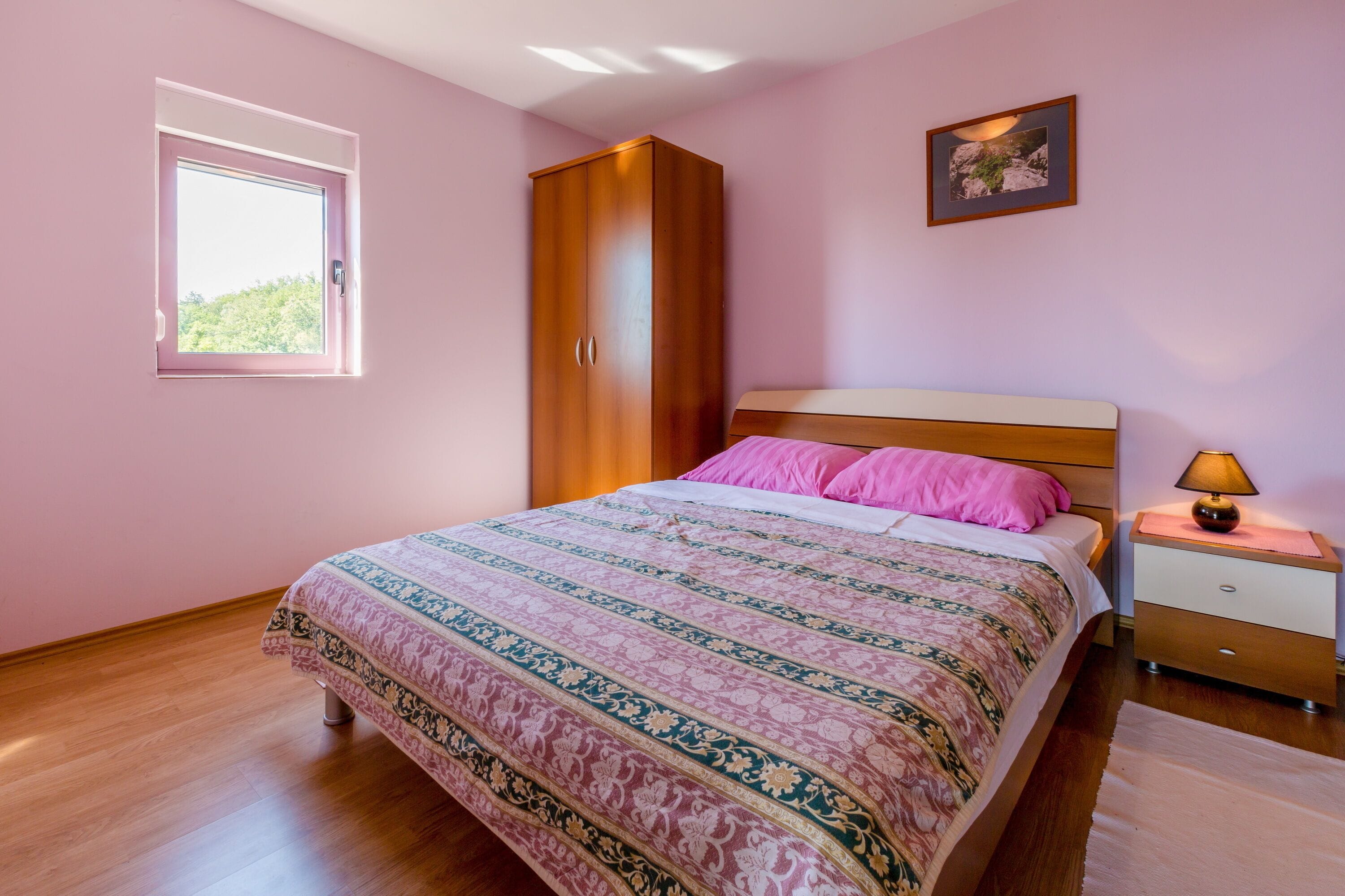 A bedroom with a bed, a wardrobe, a nightstand, a lamp, and a window.