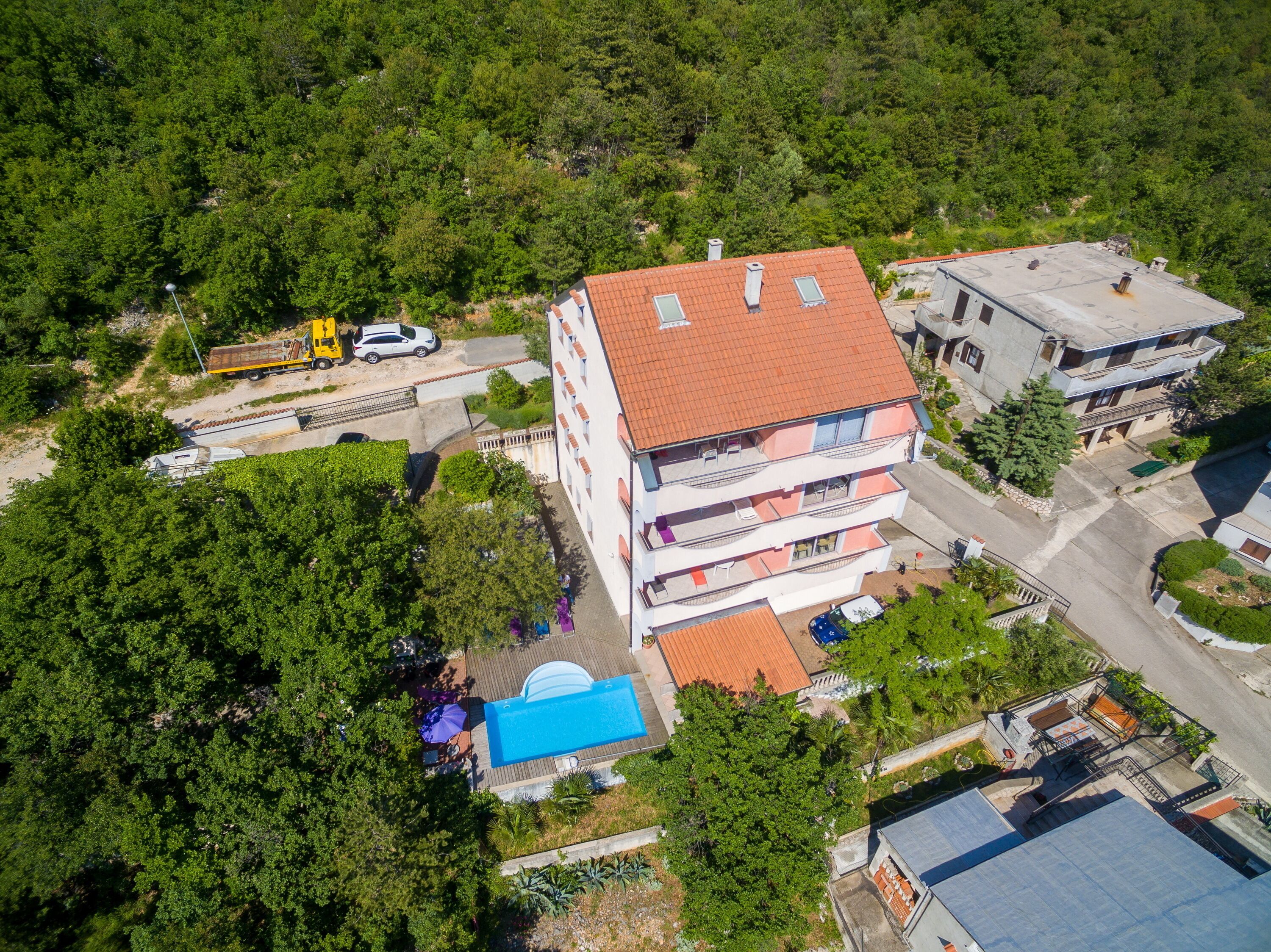 An aerial view of a multi-story building with a red-tiled roof, a swimming pool, and a parking area with a yellow truck.
