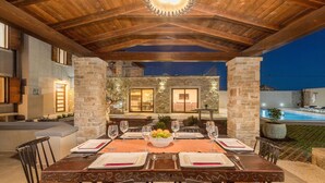 Un espacio de comedor al aire libre con pared de piedra, techo de madera y vista a una piscina.
