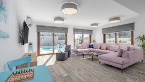 Un moderno salón con un sofá rosa, una mesa de centro y un tablero de ajedrez sobre una mesa azul. Se ve una piscina a través de las puertas de cristal.