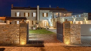 Una casa de dos pisos con fachada de piedra, entrada protegida por una reja y un camino de acceso bien iluminado.